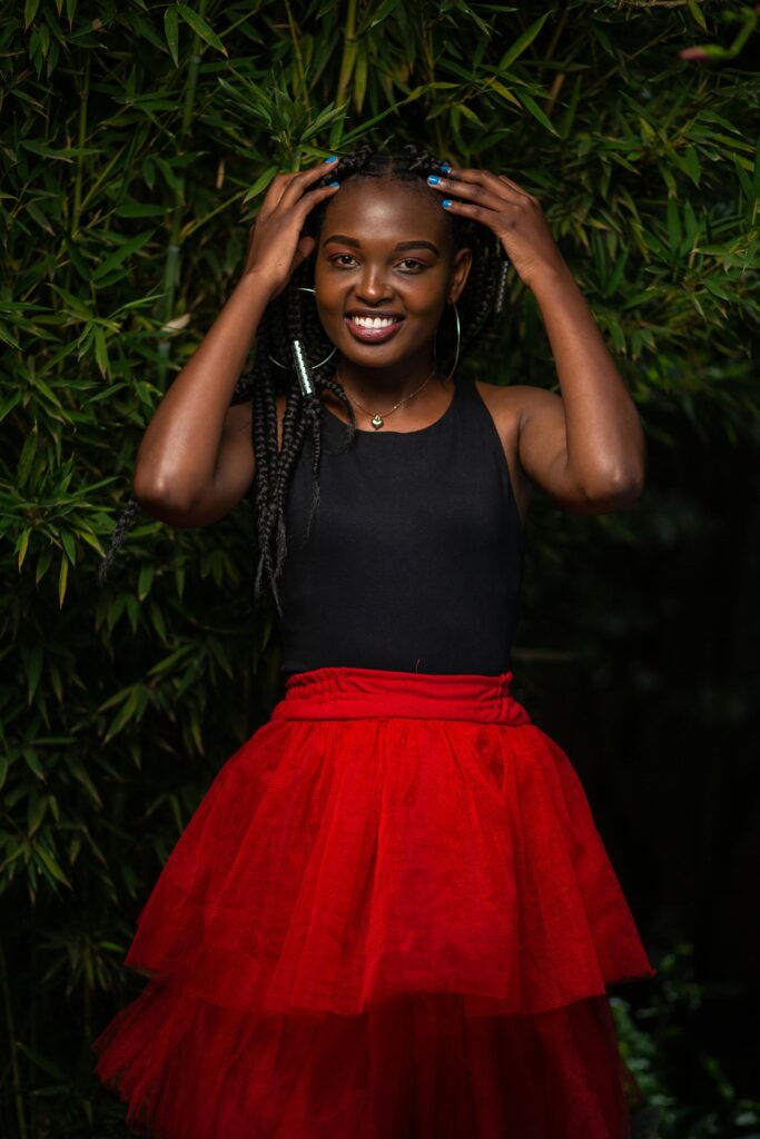 A smiling woman poses with hands on head, wearing a black top and red skirt, surrounded by greenery.