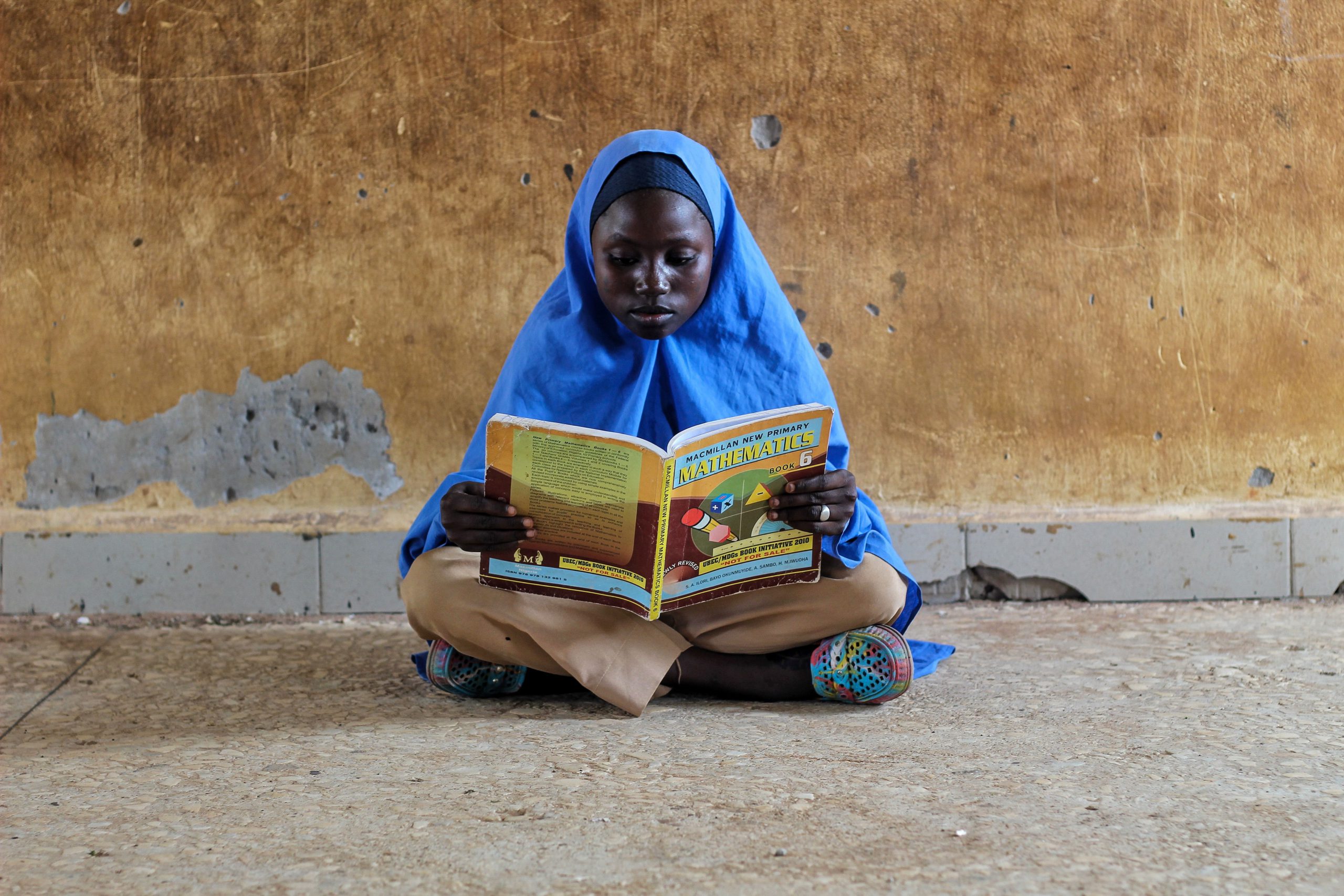 A young girl in hijab sitting on a floor reading a mathematics book intensely.